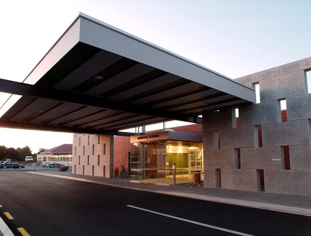 Exterior architectural photograph of a contemporary commercial building entrance at dusk. A large projecting flat canopy with dark exposed structural ribs shelters the glazed entry, while the facade combines smooth grey concrete or stone panels with narrow vertical openings and a warm-toned entry volume. A paved driveway and covered drop-off area dominate the foreground.