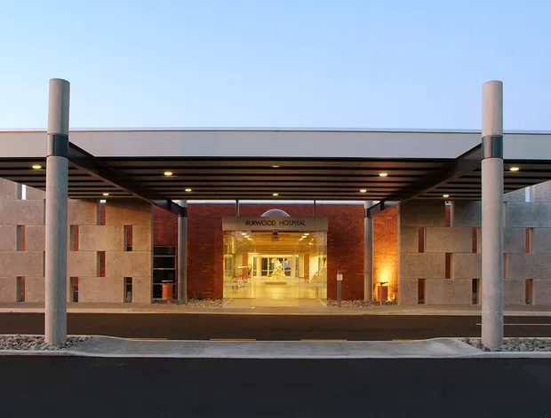 A symmetrical modern commercial building entrance viewed from outside at dusk. The broad flat-roofed canopy is supported by tall cylindrical concrete columns and shelters a recessed, warmly illuminated glazed lobby. Flanking facade walls use pale masonry or concrete panels with narrow vertical slit windows, while dark timber-toned soffit slats and red-brown brick or stone around the entrance provide contrast. A paved forecourt and roadway occupy the foreground.