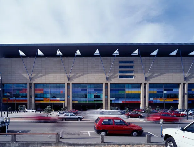 Exterior view of a large contemporary commercial or institutional building with a broad flat projecting roof, exposed triangular structural supports, pale masonry or concrete facade, and a colourful strip of glazed panels at ground level. The building fronts a busy urban street with parked and passing vehicles.