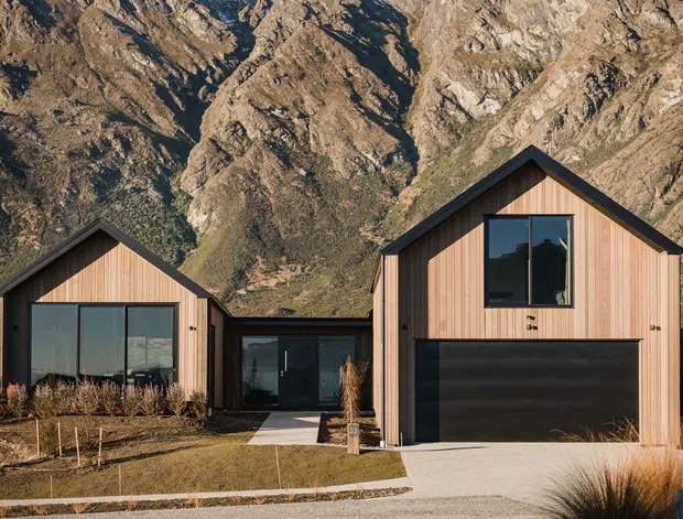 Contemporary residential development of detached gable-roof homes clad in vertical warm timber, with dark-framed glazing and dark garage doors. The houses sit against a dramatic rugged mountain backdrop, with restrained landscaping, concrete paths, and ornamental grasses.