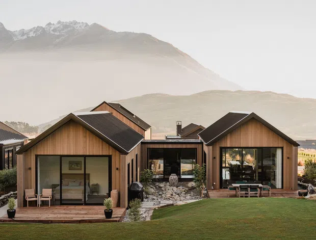 A group of compact contemporary cabins with steep gabled roofs, dark metal roofing, and warm vertical timber cladding. Large glazed openings connect the cabins to small timber decks and a landscaped lawn, while rugged mountains rise behind the development.