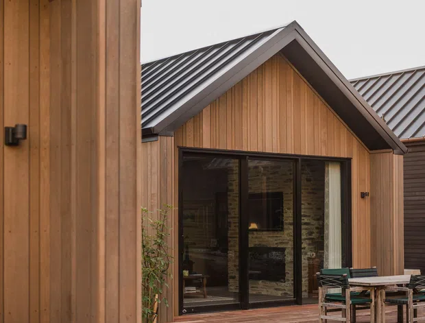 Exterior view of a contemporary timber-clad building arranged around a courtyard or outdoor dining area. Vertical warm-toned wood boards, dark metal-framed glazing and a pitched gable roof with grey standing-seam metal roofing define the facade. Outdoor tables and chairs are visible in the foreground, with another dark-clad wing to the right.