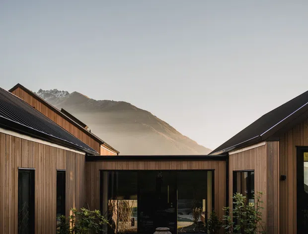 A contemporary timber-clad building arranged around a central courtyard or glazed connecting pavilion. Dark pitched roofs and vertical warm-toned timber siding frame large black-framed openings, with planted courtyard edges and a dramatic mountain landscape visible beyond.