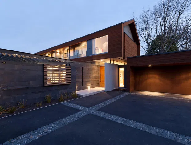 Contemporary two-storey residence viewed from the driveway at dusk, featuring dark timber upper cladding, concrete masonry walls, extensive upper-level glazing, a covered entry, and an attached double garage. The forecourt is paved in dark asphalt with contrasting bands of small grey stone setts, while warm architectural lighting illuminates the entry and garage.