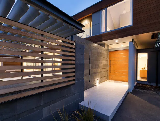 Contemporary residential entry viewed from outside, featuring a covered walkway, horizontal timber privacy screening, dark masonry walls, warm timber cladding, a recessed timber front door, and large upper-level glazing. Integrated lighting and clean geometric forms create a refined modern facade.