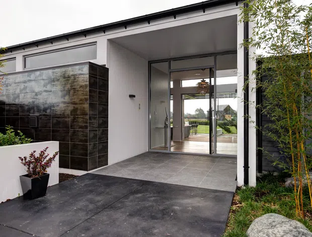 Contemporary single-storey home entrance with a covered entry, large glazed pivot or sliding doors, dark tiled feature wall, grey tiled threshold, white rendered walls, and a landscaped approach with bamboo planting.