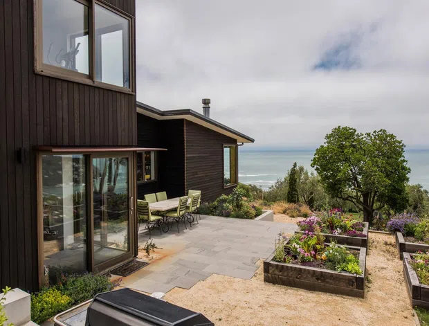 Exterior view of a contemporary dark-clad coastal home, showing large glazed openings, a paved terrace with outdoor dining, raised garden beds, and an ocean view beyond the landscaped site.