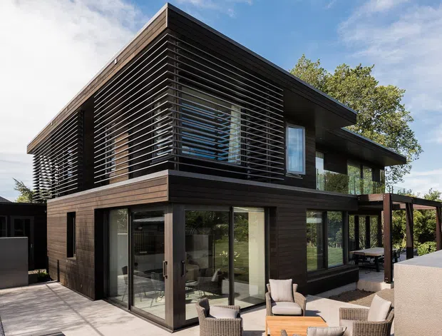 Contemporary two-storey residence with dark timber-clad walls, extensive floor-to-ceiling glazing, and a prominent upper-level horizontal louvred screen. The home opens onto a paved outdoor terrace with seating and a covered pergola-like extension, surrounded by mature landscaping.