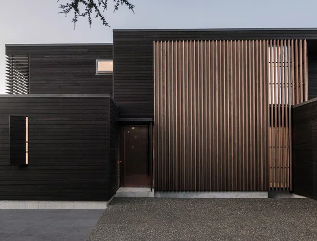 Front exterior view of a contemporary multi-level residence with dark horizontal cladding, a tall vertical timber batten screen, recessed entry door, narrow vertical window, and a textured driveway. The composition emphasizes the building facade and its layered, rectilinear volumes.