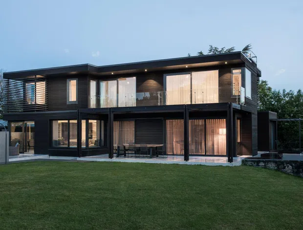 Exterior view of a contemporary two-storey residence with a strong rectilinear form, dark cladding and framing, extensive glazing, timber privacy screens, and a broad lawn in the foreground. Recessed outdoor living areas and warm interior lighting are visible beneath a flat roofline.
