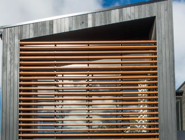 Modern building facade featuring a large recessed glazed opening screened by closely spaced horizontal timber louvres. The exterior is clad in vertically textured grey material, with strong geometric lines and a partially visible pitched roof behind the screen.