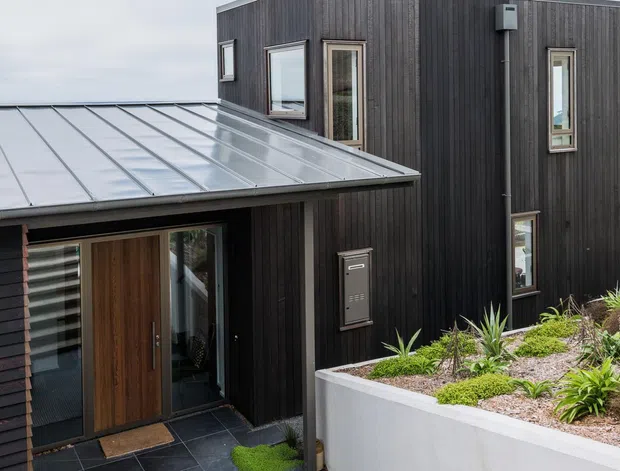 Exterior view of a contemporary multi-level residence with dark vertical timber cladding, a low-pitched standing-seam metal roof, timber entry door, narrow vertical windows, and a landscaped raised planter along the facade.