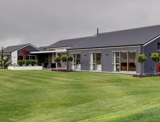 A wide exterior view of a low, single-storey residential home set behind an expansive manicured lawn. The building has dark grey horizontal cladding, multiple pitched gable roofs, white-framed glazing and doors, and a covered outdoor entertaining area with restrained landscaping. Overcast conditions create a subdued, contemporary suburban presentation.