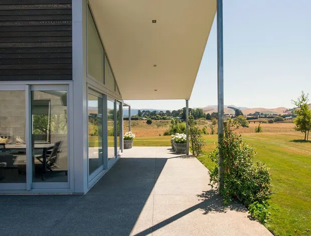 Exterior view of a contemporary, low-profile building with a projecting flat roof and covered concrete terrace or walkway. The facade combines grey masonry block, extensive floor-to-ceiling glazing, and dark horizontal louvres. Through the glazing, a furnished interior is visible. The property overlooks an expansive landscaped lawn and rural agricultural landscape.