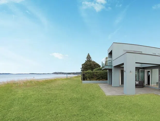 Wide exterior view of a modern, low-profile coastal home set on a large lawn beside a beach or waterfront. The building has crisp white rendered walls, broad flat roof forms, extensive glazing, covered outdoor living areas, and a minimalist geometric composition.