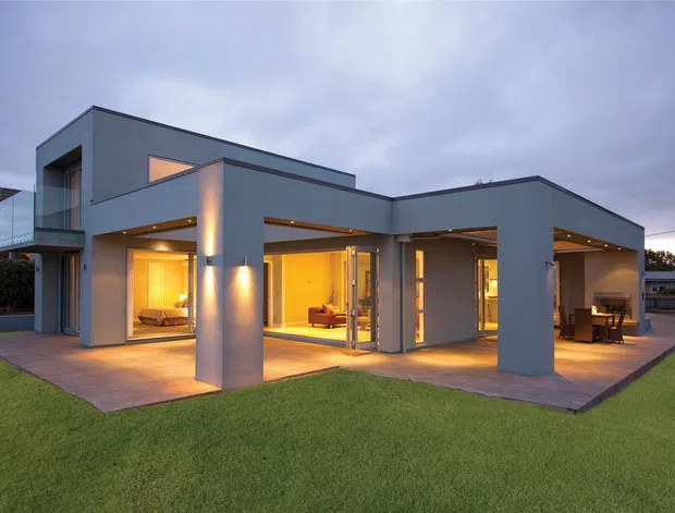 Exterior dusk view of a contemporary two-storey flat-roofed house with a strong geometric composition, smooth dark grey rendered walls, extensive floor-to-ceiling glazing, covered outdoor living areas, and warm interior lighting. A broad lawn surrounds the paved terrace, with visible outdoor dining furniture and glimpses of the living spaces inside.
