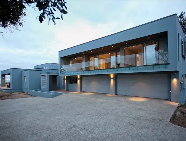 A modern multi-level residential exterior with a long rectangular form, flat roof, grey rendered walls, extensive upper-level glazing with glass balustrades, recessed downlighting, and two integrated double garages. A broad concrete driveway leads to the building, with mature trees at the edges.