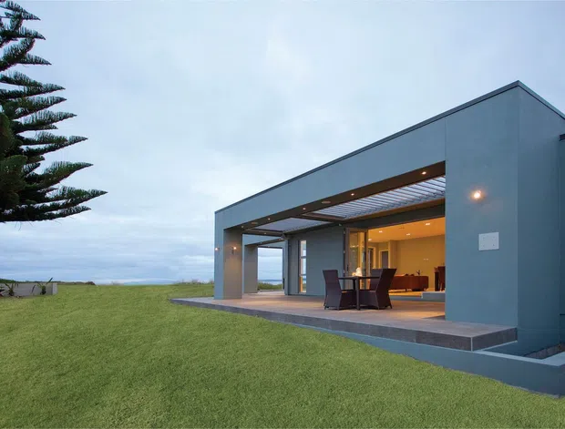 A contemporary single-storey pavilion-style house with a strong geometric form, deep covered terrace, and extensive open glazing. The muted blue-grey rendered exterior frames a warm, illuminated living area and outdoor dining setting. The home sits on a broad lawn beside a visible ocean horizon, with a large Norfolk Island pine nearby.