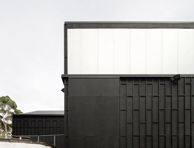 Exterior view of a stark contemporary black-clad building with a large gridded industrial-style facade, high translucent upper wall panels, and a broad paved forecourt. A low white retaining wall and partial trees sit at the edge of the site.