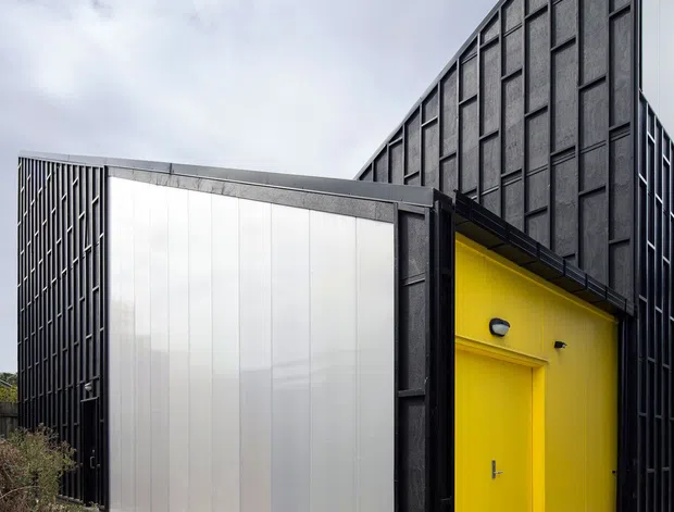 Exterior architectural photograph of a sharply angular, contemporary building with a tall triangular gable-like form. The facade combines dark framed textured panels and translucent corrugated cladding, with a vivid yellow recessed entry door creating a strong accent. Concrete paths and gravel landscaping surround the structure under an overcast sky.
