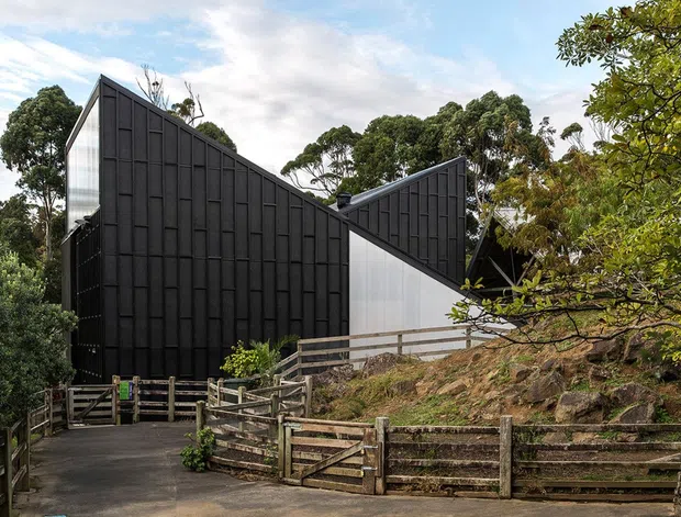 Exterior view of a contemporary dark-clad building set within a wooded, sloping site. The structure has sharply angled, folded rooflines and facade planes, with black vertical panel cladding contrasted by a light translucent or smooth wall section. Timber post-and-rail fencing, a paved approach, exposed rock, and mature trees surround the building.
