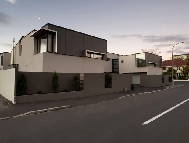 Exterior street view of a contemporary multi-level residential development composed of two angular homes. The buildings feature flat rooflines, crisp geometric volumes, dark vertical upper cladding, smooth light rendered walls, recessed balconies and large dark-framed windows. Low boundary walls and minimalist planting line the paved street frontage.