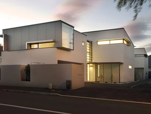 Street-facing view of a contemporary multi-storey residential building at dusk, composed of crisp rectangular volumes with smooth pale walls, dark grey upper cladding, projecting box forms, narrow horizontal windows, extensive vertical glazing, and external louvres. The building sits within a dense urban or suburban setting with a driveway and neighbouring structures visible.