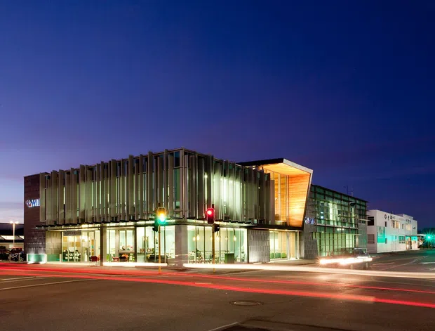 Night-time exterior view of a contemporary multi-storey commercial building on an urban street corner. The facade combines extensive floor-to-ceiling glazing with vertical fins, a projecting warm timber-lined upper volume, and a long glazed side wing. Traffic lights, road markings, and light trails emphasize the prominent street-facing urban setting.