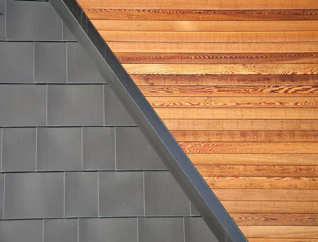 Close-up architectural detail showing a diagonal junction between dark grey rectangular exterior cladding tiles and a warm-toned timber-lined surface. The timber has visible horizontal boards with pronounced natural grain and layered trim detailing, creating a strong geometric contrast.