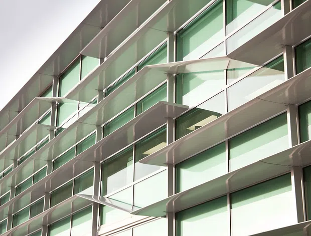 Close-up exterior view of a contemporary multi-storey building facade with extensive green-tinted glazing, silver metal framing, and projecting horizontal balcony or sunshade elements. The repetitive geometric composition emphasizes modern facade design and solar shading.