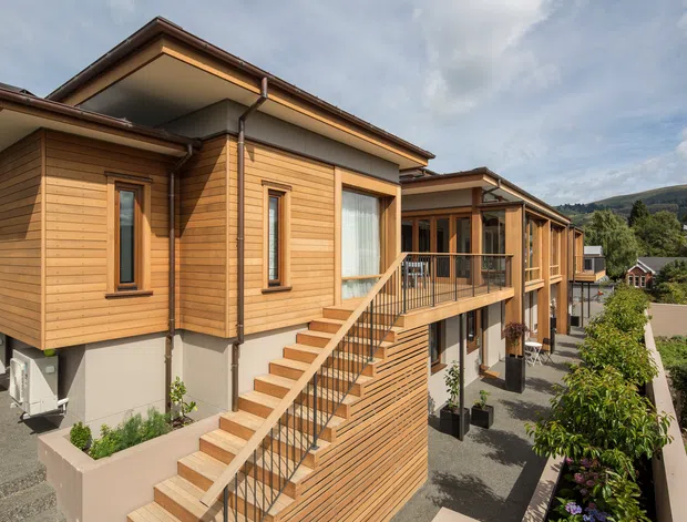 Exterior view of a contemporary multi-level residential building clad primarily in warm horizontal timber weatherboards. The home has projecting upper volumes, sloped roof planes with broad eaves, large windows, an external timber stair, and a raised balcony with dark metal balustrades. A landscaped courtyard and neighbouring hillside homes are visible in the background.