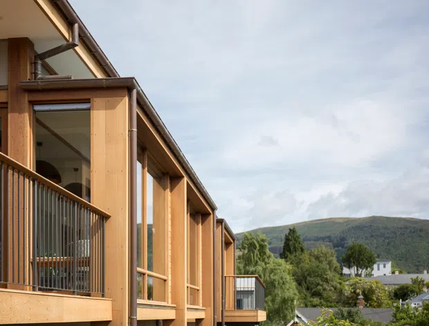 Exterior view of a contemporary timber-clad multi-level building with projecting balconies, large glazed openings, exposed vertical timber posts, and dark metal gutters and balustrades. A green, hilly landscape and neighbouring homes are visible beyond the building.