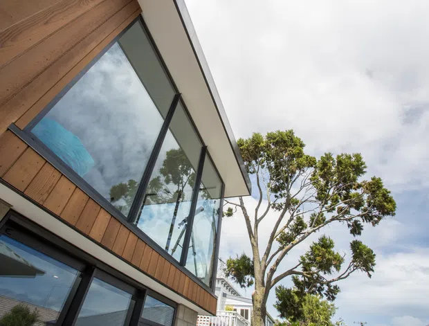 Low-angle exterior view of a contemporary multi-level building with warm timber cladding, dark-framed expansive glazing, and a projecting roof or overhang. A glazed upper-level volume and balcony railing are visible, with mature planting beside the building.