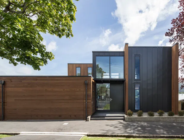 Street-facing exterior of a contemporary multi-volume home, featuring a broad horizontal timber-clad garage or living wing beside a taller dark vertically clad section. Large glazed entry and upper-level windows create a strong contrast between warm timber and charcoal metal cladding. A paved frontage, clipped shrubs, mature trees, and blue sky complete the composition.