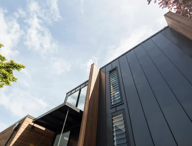Low-angle exterior view of a contemporary multi-level residential building. The facade combines dark vertically seamed cladding with warm timber soffits and projecting timber elements, narrow louvred windows, and a glazed balcony or upper-level opening. A rain chain hangs beneath the overhang, with sky and surrounding foliage in the background.