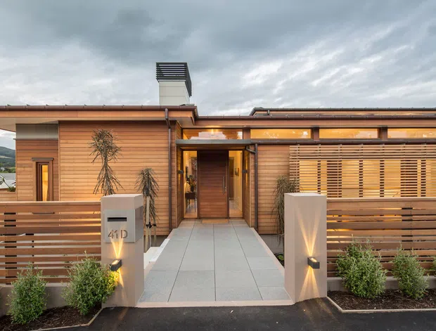 Front exterior view of a low-rise contemporary residence with warm horizontal timber cladding, a shallow roofline, deep eaves, and a sheltered entry walkway. Horizontal timber screening forms the front boundary and partially conceals the glazing, while illuminated rendered entry pillars and landscaped shrubs frame the approach. Overcast sky and distant hills are visible in the background.