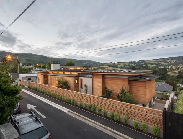 Exterior view of a contemporary single-storey residence on a sloping suburban site, featuring warm horizontal timber cladding, low-pitched metal roofing, broad roof overhangs, and a timber privacy fence. The home overlooks a green valley with hills and scattered suburban development. A paved street and overhead utility lines are visible in the foreground.