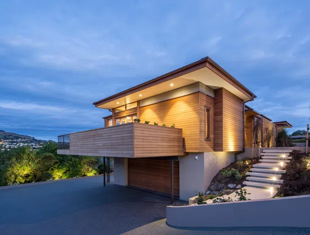 Twilight exterior view of a contemporary multi-level house on a sloping site. The home features warm horizontal timber cladding, broad roof overhangs, recessed exterior lighting, a projecting balcony, an integrated garage, and illuminated steps leading up through landscaped planting. A suburban hillside setting is visible in the background.