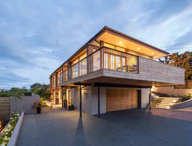 Twilight exterior view of a contemporary multi-level home on a sloped site. The building features a projecting upper-floor terrace with black metal balustrades, horizontal timber-look cladding, warm recessed lighting, large glazed openings, and an integrated double garage with a timber garage door. Landscaped stepped access and paved driveway surround the house.