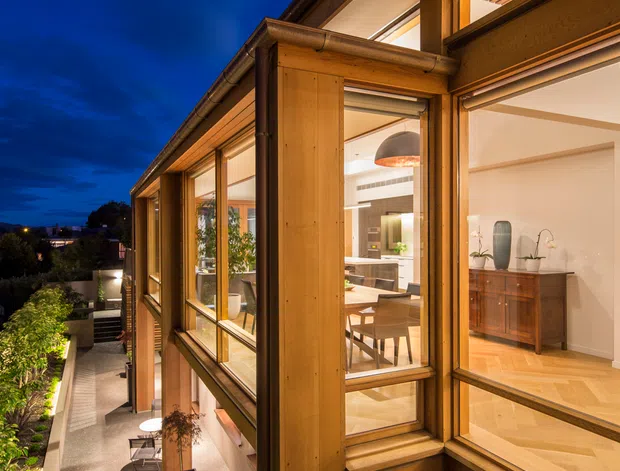 A dusk exterior view of a contemporary multi-level residence with extensive timber-framed glazing. Warm interior lighting reveals a dining area, kitchen, and living spaces through the windows, while a narrow landscaped terrace and walkway run alongside the facade. The building features projecting eaves, exposed timber framing, large windows, and a warm natural-material palette against a deep blue evening sky.