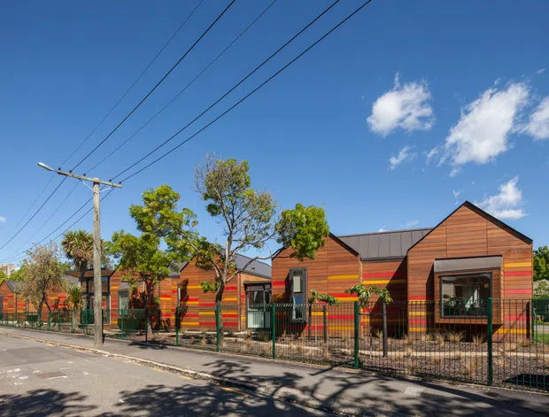 Street-level exterior view of a low-rise contemporary building composed of several small gabled volumes. The facade uses horizontal warm timber-toned cladding with vivid multicoloured horizontal accent bands, dark metal roofing, and dark-framed windows and doors. A metal security fence, paved street, utility poles, and mature trees are visible in front of the building under a clear blue sky.