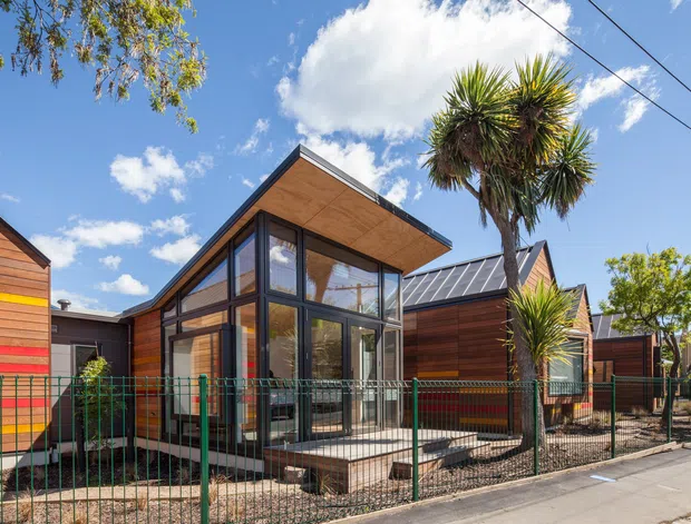 Exterior view of a contemporary timber-clad residential building with a prominent glazed corner extension, broad timber soffit, dark metal-framed glazing, and pitched metal-roofed volumes. A fenced frontage, paved path, established planting, and clear sunny sky are visible.