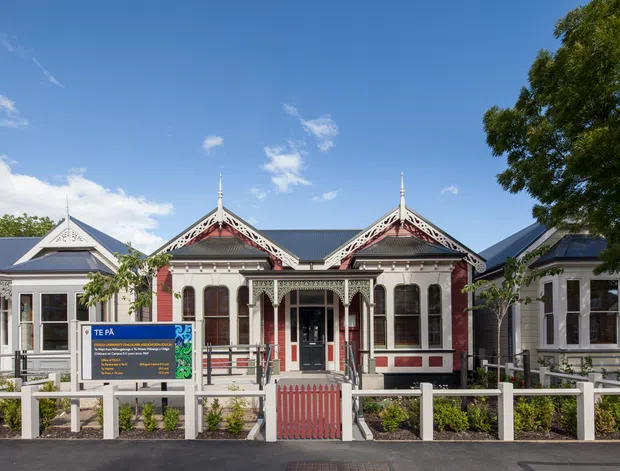 Front-facing exterior view of a historic single-storey building with ornate Victorian-era detailing. The symmetrical facade features decorative gable bargeboards, steep pitched roofs, white weatherboard cladding, red and dark trim, tall multi-pane windows, and a covered central entry porch. A sign and landscaped frontage sit behind a low white fence.
