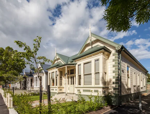 Exterior view of a restored ornate single-storey timber villa, featuring cream weatherboard cladding, decorative gable detailing, steep green metal roofs, bay windows, verandah-style entry, and a landscaped front garden along a paved urban walkway.