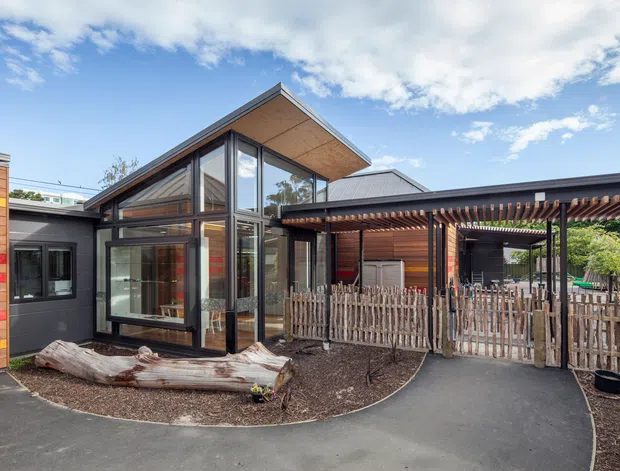 Exterior view of a contemporary single-storey building with a prominent glazed gable pavilion, dark window framing, warm timber soffits and horizontal timber cladding. A covered pergola or carport-like structure extends across the front, with a paved walkway, fenced outdoor area and landscaped garden beds.