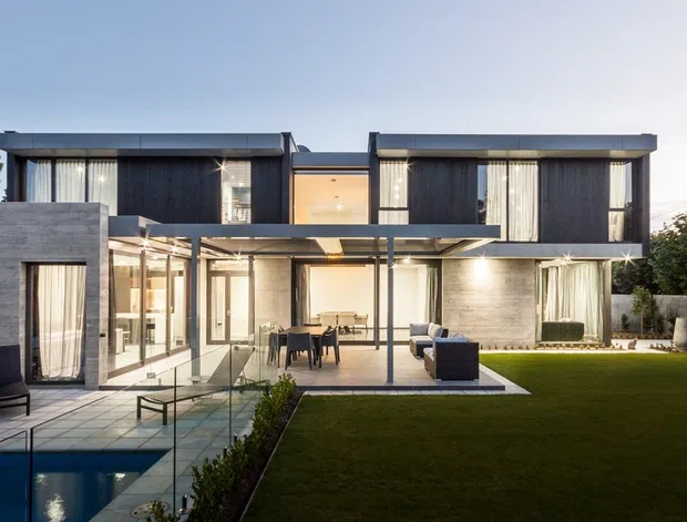 A contemporary two-storey residence with a strong rectilinear composition, dark upper-level cladding, extensive floor-to-ceiling glazing, and exposed concrete or grey masonry walls. A covered outdoor entertaining area opens onto a manicured lawn, with a narrow swimming pool and paved terrace in the foreground. The home has flat roof planes, slender dark frames, and a restrained indoor-outdoor connection.