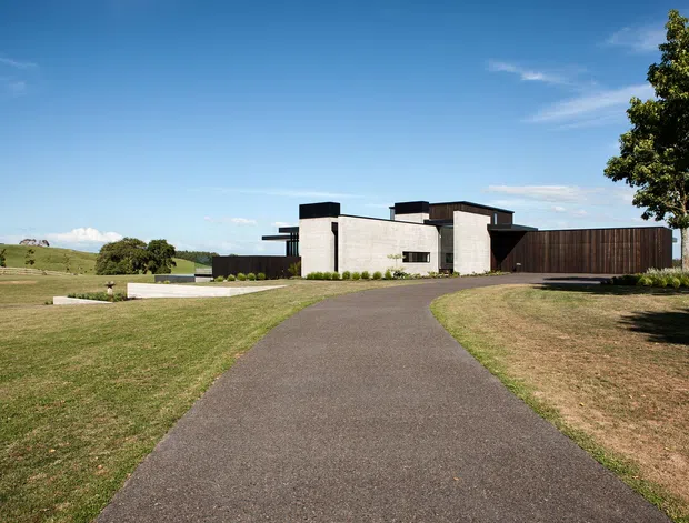 Wide exterior view of a low-profile contemporary residence set in an expansive rural landscape. The house combines pale masonry volumes, dark flat-roof elements, extensive dark timber or metal screening, and clean rectilinear forms. A paved driveway leads toward the entrance, with lawns, sparse landscaping, and open farmland surrounding the property under a clear blue sky.
