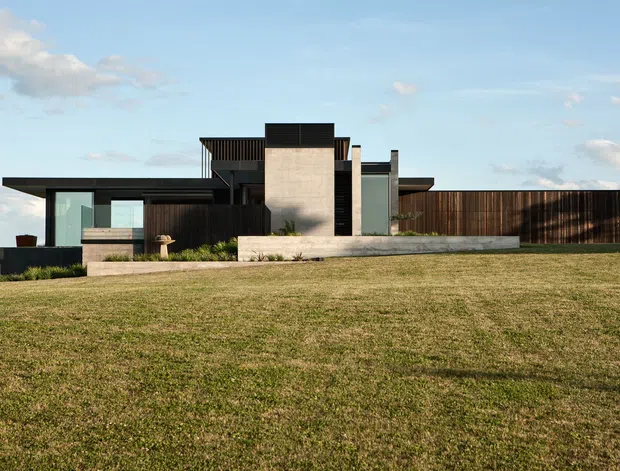 Low, horizontally composed contemporary residence set behind a broad lawn. The building features flat roof planes, dark timber-clad volumes, extensive glazing, a prominent pale concrete or rendered central block, and long linear retaining walls and boundary elements. The facade is viewed from the exterior under a clear sky, with simple landscaped planting around the entry terrace.