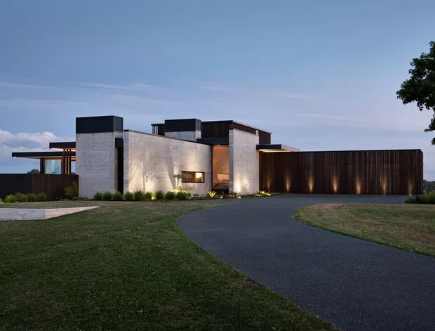 A low, modern residence photographed at dusk, composed of interlocking rectilinear volumes. Pale concrete or rendered masonry walls contrast with dark timber-clad upper volumes and a long dark garage-like wing. A broad paved driveway curves through the lawn toward the house, with subtle exterior lighting and sparse planted borders. The flat-roofed composition and strong horizontal and vertical geometry create a restrained contemporary exterior.