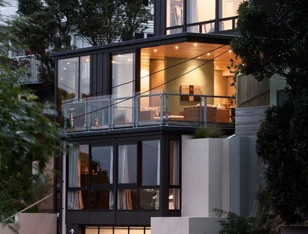 Vertical dusk exterior view of a multi-level urban residence on a sloped site. The house combines a white weatherboard upper storey with a dark, contemporary glazed extension featuring cantilevered volumes, extensive black-framed windows, warm recessed lighting, a balcony with glass balustrade, and an integrated illuminated garage door. Rendered retaining walls, stepped levels, and dense planting frame the facade.